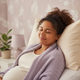 Headshot of a happy, pregnant woman, smiling contentedly while resting on a pillow, with warm cream and soft lavender blanket
